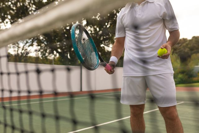 Low section of Caucasian man wearing tennis whites spending time on a court playing tennis on a sunny day, holding a tennis racket and a ball, seen through net. Hobby sport leisure time.