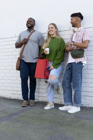 Multiracial friends leaning on white brick wall holding coffee, shopping bag and phone