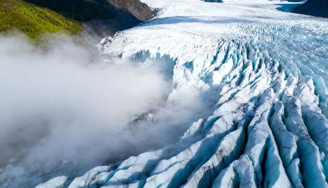 Flowing alpine glacier tongue with deep crevasses and mist over rugged mountain valley expanse