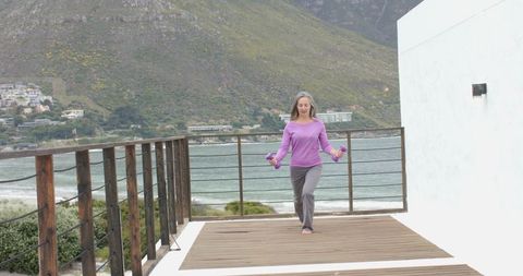 Mature woman lunging on sea deck holding purple dumbbells barefoot overlooking bay