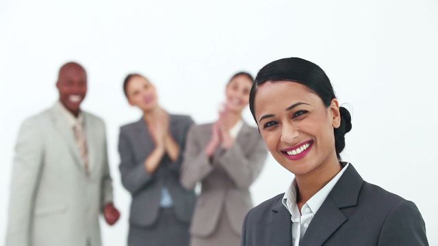 Businesswoman smiling while receiving applause from her colleagues. Colleagues stand in the background clapping and showing their support. Ideal for depicting concepts of teamwork, appreciation in the workplace, diversity in business environments, and achievement recognition.