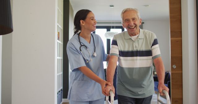In the image, a cheerful elderly man using a walker is receiving support from a caregiver in a home environment. The caregiver, dressed in scrubs with a stethoscope around her neck, is helping and encouraging the senior during his rehabilitation. This can be used to represent themes like aging in place, senior support, assisted living, healthcare, nursing home promotion, and depicting the compassion of healthcare workers.