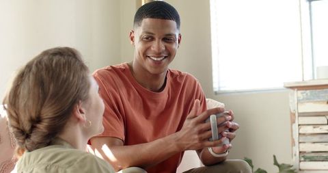 Smiling young man holding mug while chatting with friend in sunlit modern living room