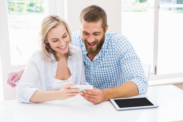Couple sitting at a table using a smartphone, smiling and enjoying their time together. Tablet on the table suggests they are tech-savvy. Ideal for use in advertisements for technology, communication services, or lifestyle blogs focusing on modern relationships and home life.