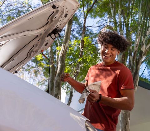 Young man checking car oil and smiling in driveway under trees during sunny afternoon