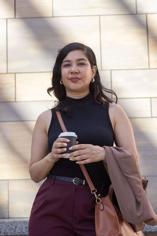 Urban woman standing holding takeaway coffee in black sleeveless top and maroon trousers