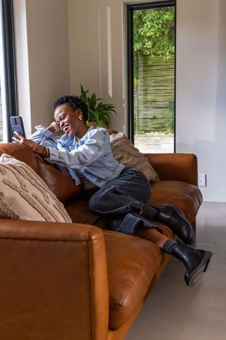 Smiling african american woman relaxing on caramel leather sofa with smartphone in sunlit lounge