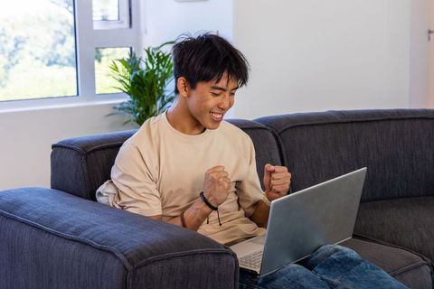 Young asian man celebrating with laptop on sofa at home enjoying remote success smiling