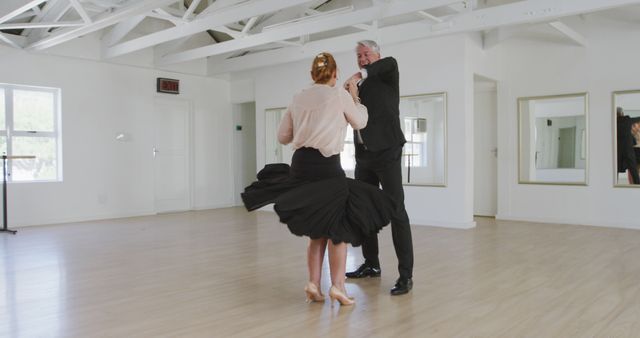 Senior couple gracefully dancing in a bright dance studio with mirrors on the wall. The man wears a dark suit, while the woman wears a light blouse and a flowing black skirt. This can be used for themes related to active aging, dance classes, hobbies, and romantic relationships in later life.