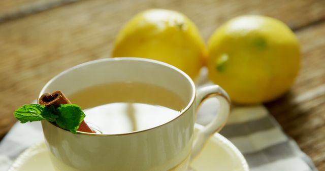 A cup of tea with a cinnamon stick and mint leaf garnish sits on a table, accompanied by fresh lemons in the background. The warm beverage suggests a cozy setting for relaxation or a home remedy for wellness.