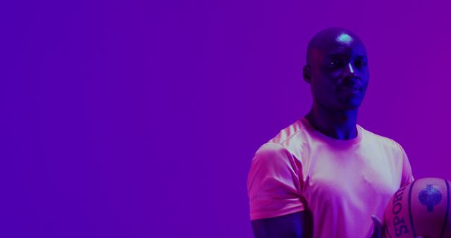 A black man wearing a light athletic shirt holds a basketball while standing in vivid purple neon lighting. Ideal for advertisements about sportswear, fitness programs, motivational posters or gym promotions.