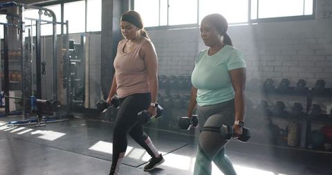 Two diverse women performing weighted lunges with hex dumbbells in bright gym