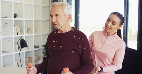 Senior man doing dumbbell exercises at home while caregiver guiding gentle strength rehab