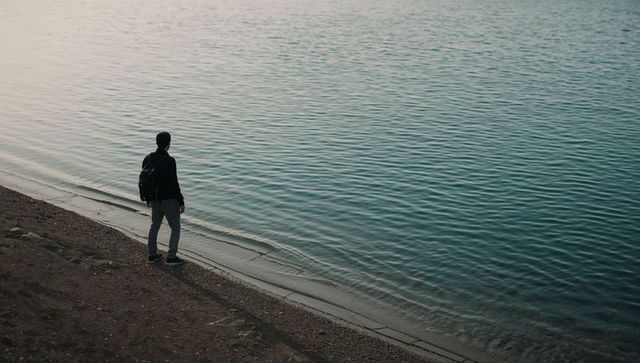 Solitary man standing at lakeside facing rippling water on pebble shore, overcast calm