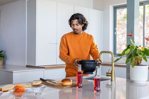 Indian man preparing burger in modern kitchen wearing orange sweater holding colander over sink