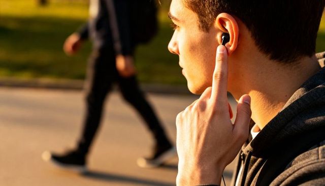 Teenage male pressing in-ear earbud while listening on sunlit park walkway at golden hour