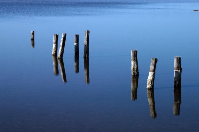 Image show remnant pilings from old dock reflected in calm, blue waters near Kennedy Space Center. Captures serene and peaceful atmosphere. Ideal for use in nature conservation materials, environmental awareness campaigns, landscape photography collections, and tranquility-themed designs.