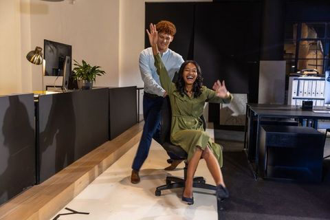 Diverse coworkers enjoying playful rolling chair race through modern creative workspace