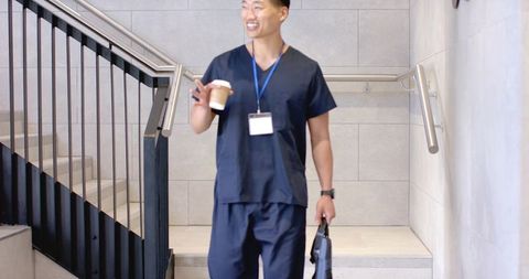 Asian healthcare worker walking down clinic stairwell holding coffee and bag, wearing navy scrubs