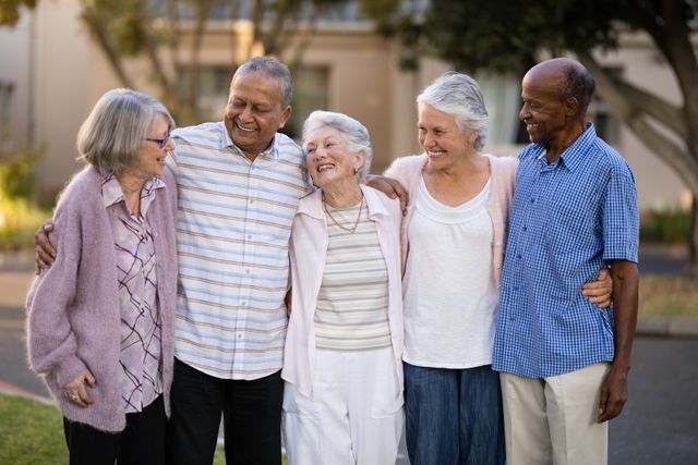 Group of senior friends standing together outdoors, smiling and embracing each other. Ideal for use in content related to senior living, retirement communities, elderly care, friendship among seniors, and promoting a positive lifestyle for the elderly.