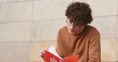 Curly-haired adult man wearing glasses reading orange spiral notebook by beige tiled wall