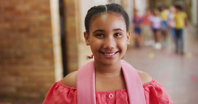 A girl wearing a red blouse and a pink scarf stands indoors, smiling confidently at the camera. She is in a school corridor with other children visible in the background, exuding a sense of joy and cheerfulness. Perfect for use in educational materials, websites focused on childhood and education, or promotional content for schools and extracurricular activities.