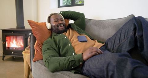 African American man relaxing on couch with headphones and smartphone by cozy fireplace