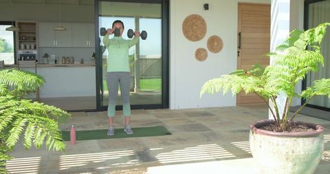 Asian woman lifting dumbbells on sunlit modern home patio with yoga mat and water bottle