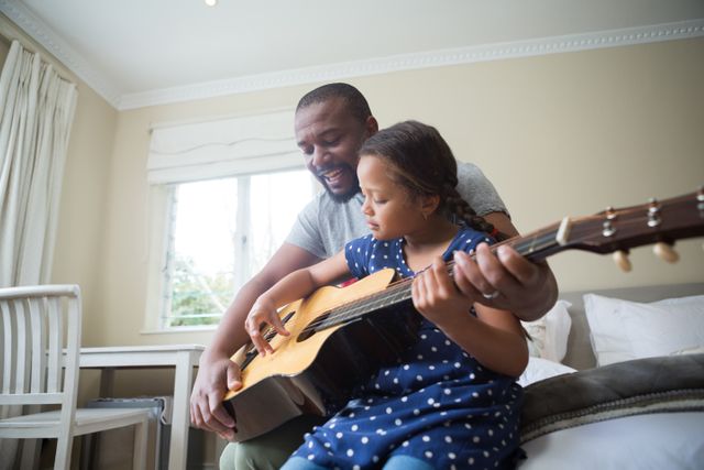 Father teaching his daughter to play the guitar in a cozy bedroom. Ideal for use in parenting blogs, family bonding articles, music education websites, and advertisements promoting family activities and learning at home.