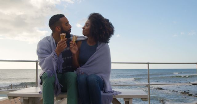 Young couple enjoying ice cream by the beach on a chilly day, wrapped in a cozy blanket. This can be used in content related to romance, leisure, seaside holidays, relationship bonding, and fun outdoor activities despite the weather.