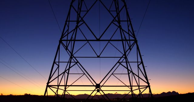 Silhouetted against a twilight sky, a power transmission tower stands tall, with copy space. Its geometric structure contrasts with the natural gradient of dawn or dusk, symbolizing the intersection of human engineering and nature.