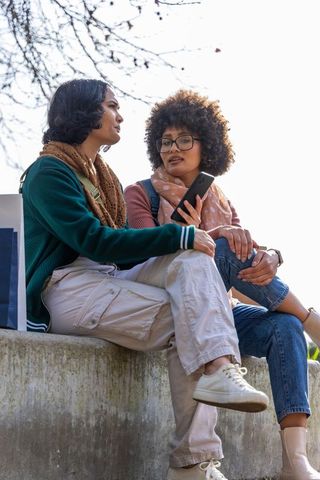 Two friends sitting on ledge using smartphone outdoors planning weekend together