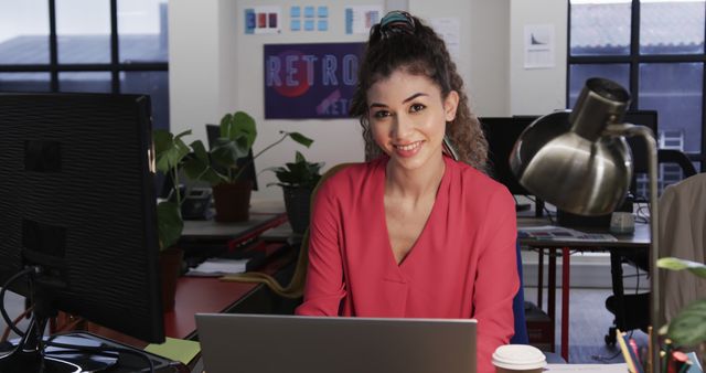 Young professional woman sits at desk in modern office, smiling while working on laptop. Casual pink blouse and relaxed atmosphere suggest creativity and productivity. Perfect for use in business, technology, and workplace culture contexts, illustrating themes of productivity, professional settings, and modern office environments.