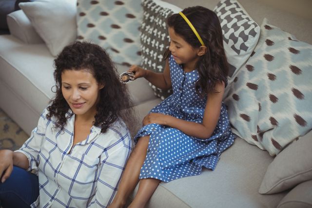 Cute daughter combing mothers hair in living room at home
