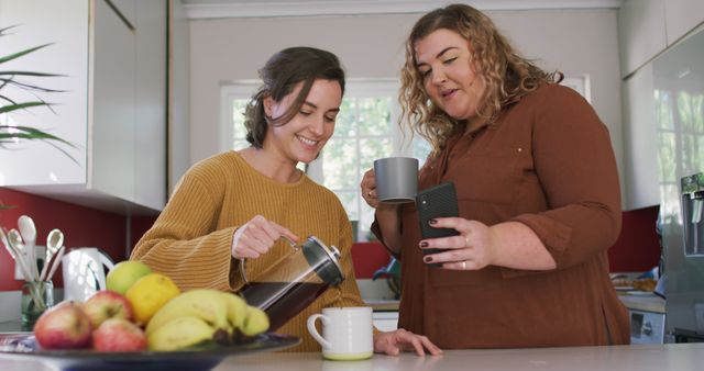 Two diverse women smiling while preparing and enjoying coffee together in a modern kitchen. One woman is pouring coffee from a French press, while the other holds a smartphone, engaging in friendly conversation. Vibrant fruits are visible on the counter, suggesting a healthy lifestyle. Ideal for use in advertisements focused on home lifestyle, kitchen appliances, friendship, or morning routines.