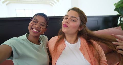 Two friends taking selfie on sofa at home, smiling and posing playfully with smartphone