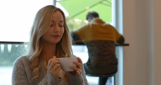 A woman is enjoying a hot beverage while sitting in a cozy cafe. The surroundings have a calm and serene atmosphere, with natural light streaming through large windows. This can be used for promoting cafes, relaxation, coffee brands, lifestyle blogs, or wellness content.