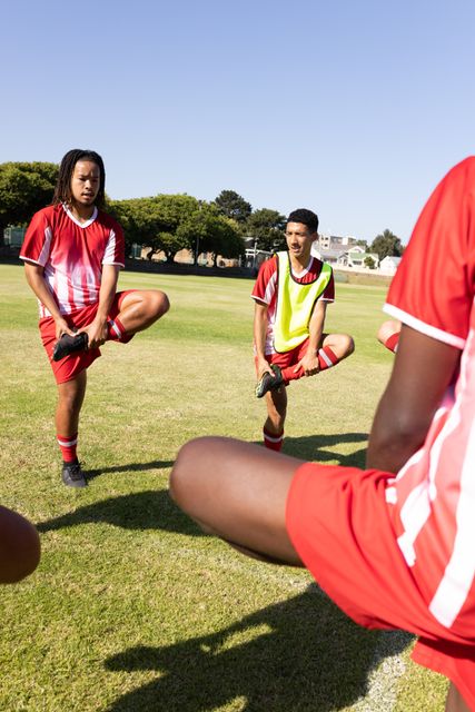 Multiracial soccer players stretching legs while standing on one leg on a grassy field under a clear sky. Ideal for use in sports and fitness articles, team-building promotions, and advertisements for athletic gear or summer sports programs.