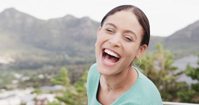 This image features a joyful woman laughing outdoors with a scenic mountain background. She is wearing a casual outfit and hoop earrings, adding a touch of everyday style to the scene. Ideal for use in campaigns promoting happiness, outdoor activities, fashion, travel, lifestyle, and health and wellness.
