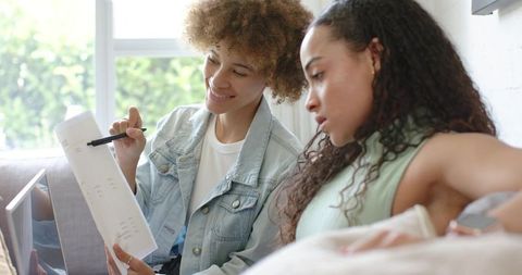 African american women collaborating on study notes with laptop and documents on sofa