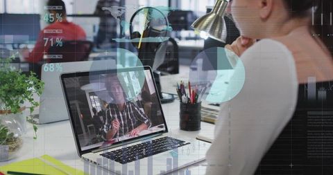 Woman watching remote video call on laptop in modern open office with analytics HUD