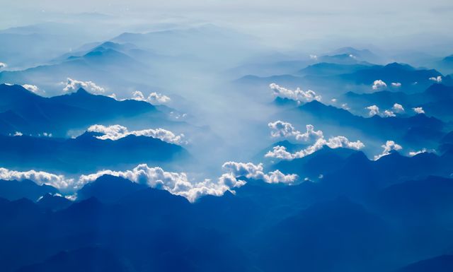 Aerial view of vast mountain range with clouds and mist blanketing the landscape. The blue hues and serene atmosphere make it perfect for use in nature-related articles, travel blogs, wallpapers, or posters. Ideal for emphasizing beauty and tranquility of natural environments.