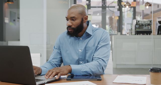 Young African American businessman working on laptop at office desk with documents and cup of coffee nearby. Perfect for depicting office work, professional environments, business productivity, and modern corporate settings in marketing materials, blogs, and website content.