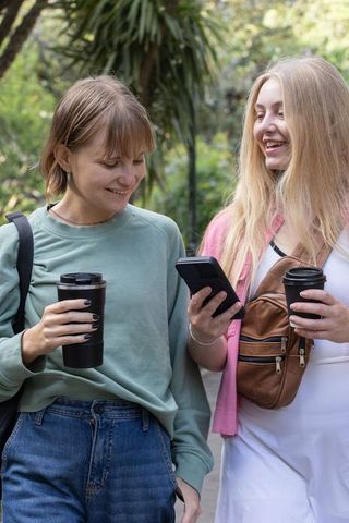 Friends walking in park checking smartphone while holding coffee travel mugs