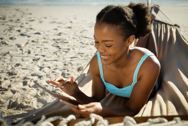 Smiling african american woman sitting in hammock using tablet on sunny the beach by the sea. holiday, romance and leisure time at the beach.