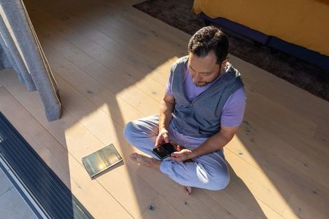 Man sitting cross-legged using smartphone on sunlit pale wooden bedroom floor