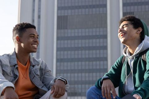 Two teenage friends laughing on urban plaza ledge with backpacks and city tower backdrop