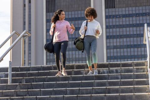 Friends walking down city steps carrying gym bags and coffee, chatting after workout