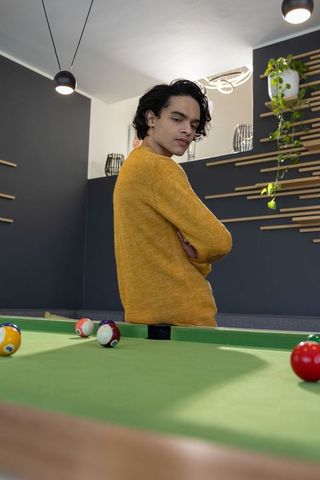 Asian man in mustard sweater looking back over shoulder at pool table in modern lounge