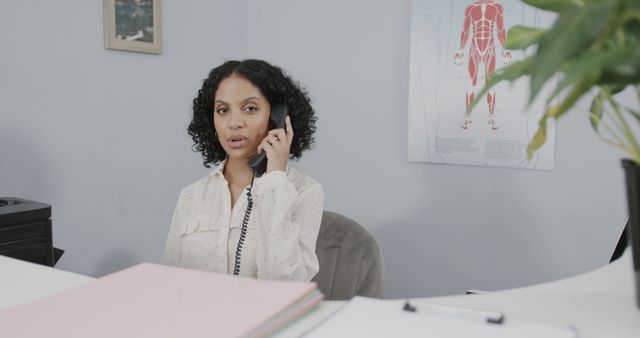 Biracial female receptionist talking on telephone at health clinic reception desk, copy space. Medical services and healthcare, unaltered.